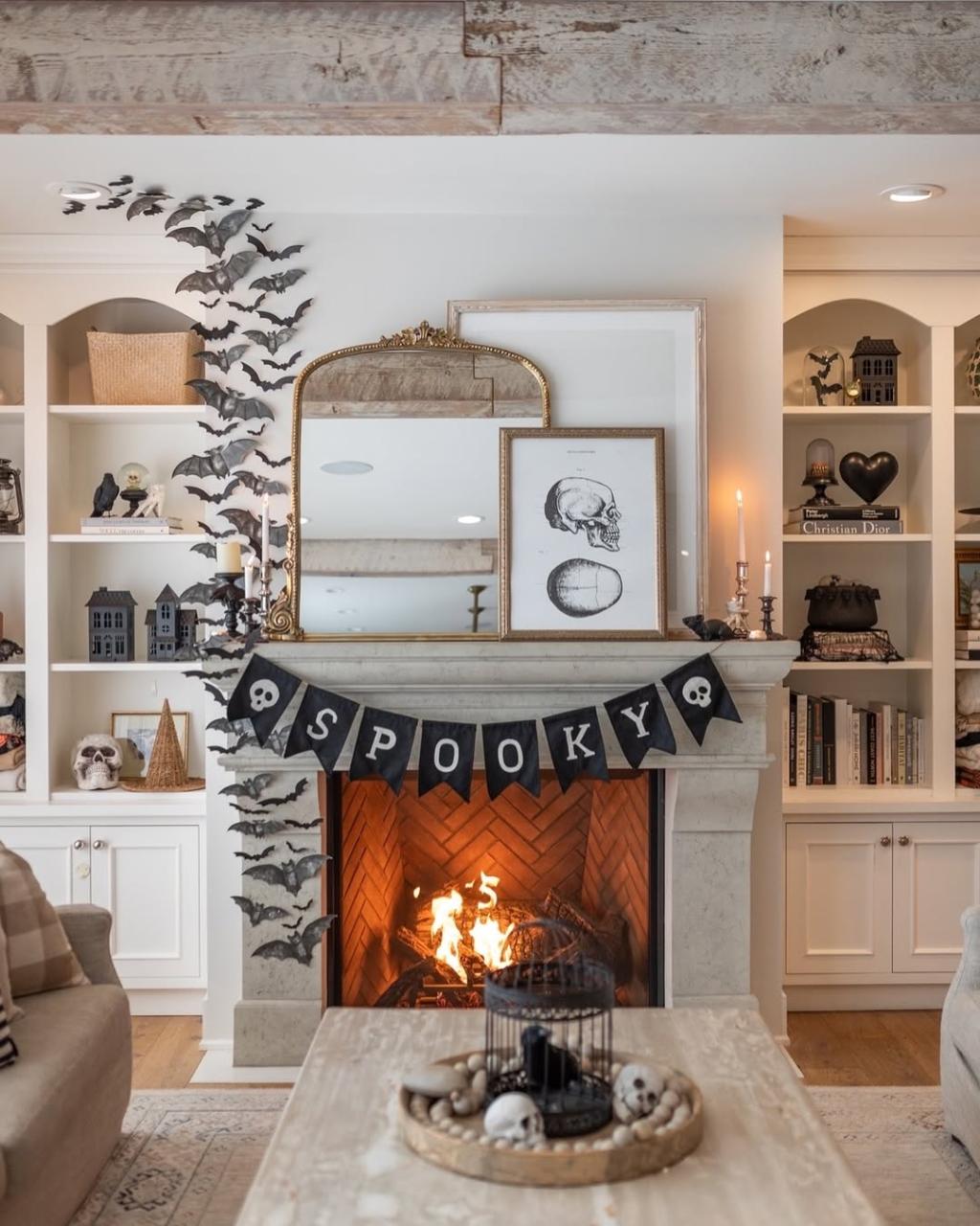 A fireplace decorated for Halloween with a "SPOOKY" banner, paper bats, skull artwork, candles, and a fire burning in the hearth—perfect inspiration for minimalist Halloween decor with bookshelves in the background.